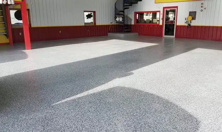 A spacious room with shiny, speckled gray flooring, red and yellow walls, windows, a glass door, and a spiral staircase leading upstairs.