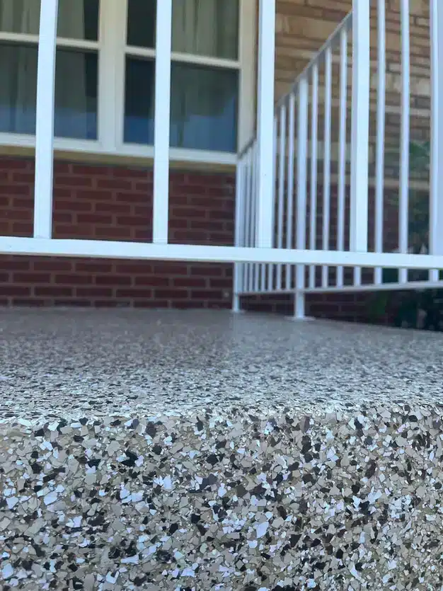 Close-up of a speckled concrete step with a white metal railing, in front of a brick facade and window with curtains.