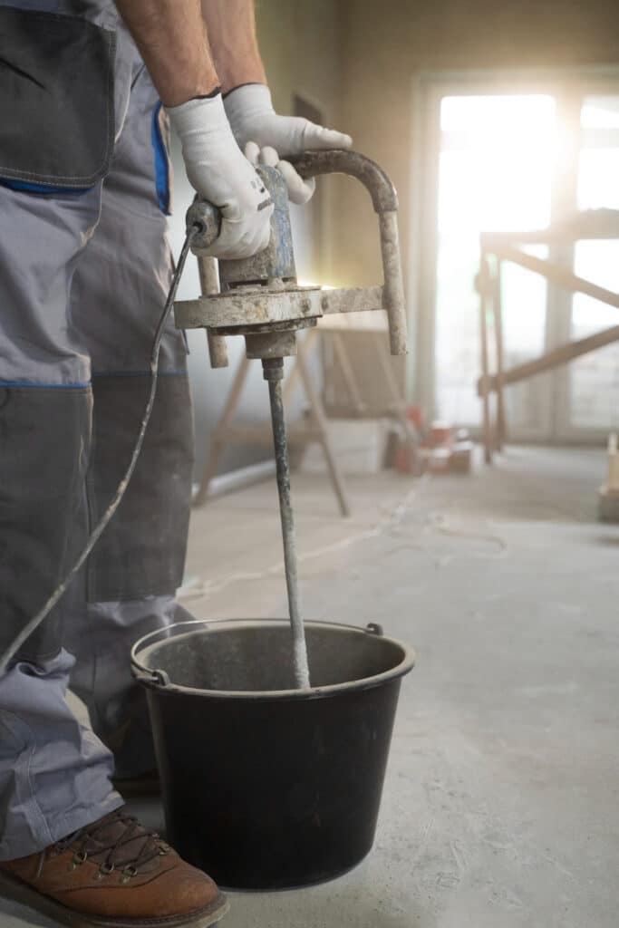 A person operates a handheld concrete mixer, stirring contents in a bucket inside a construction site with sunlit windows in the background.
