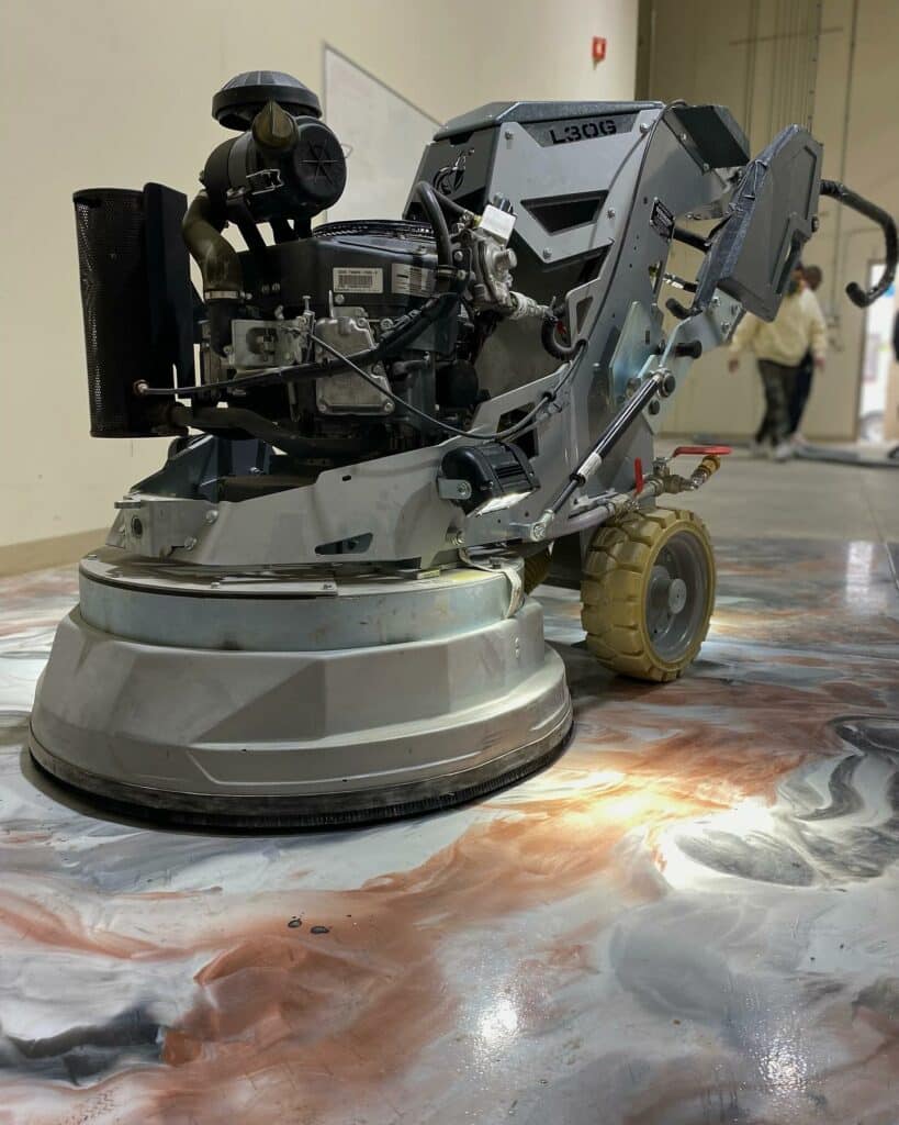 A concrete grinder machine working on a polished, marbled floor in an indoor setting. A person stands blurred in the background.