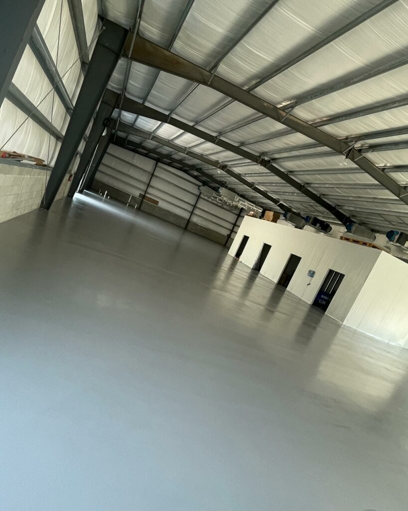 Wide-angle view of an empty, modern warehouse with a polished concrete floor, high metal ceiling, and partitioned office space. No recognizable landmarks or historical buildings.