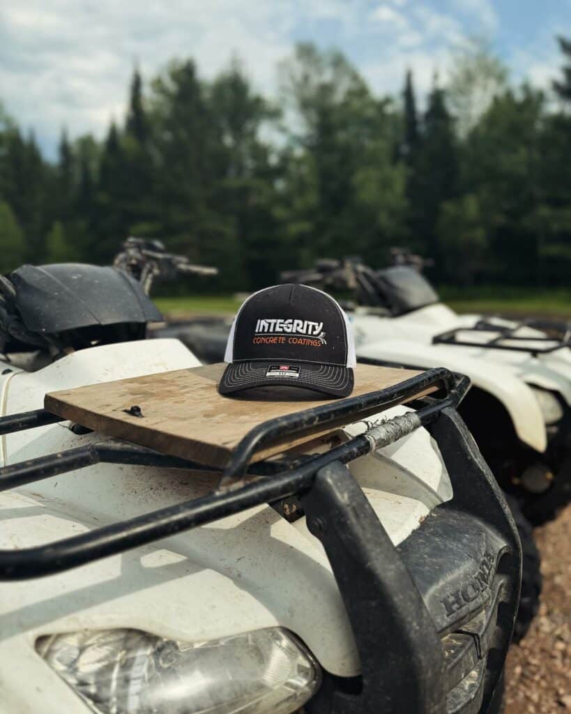 A cap with "Integrity Concrete Coatings" sits on an ATV in a forested area on a clear day; other ATVs are in the background.