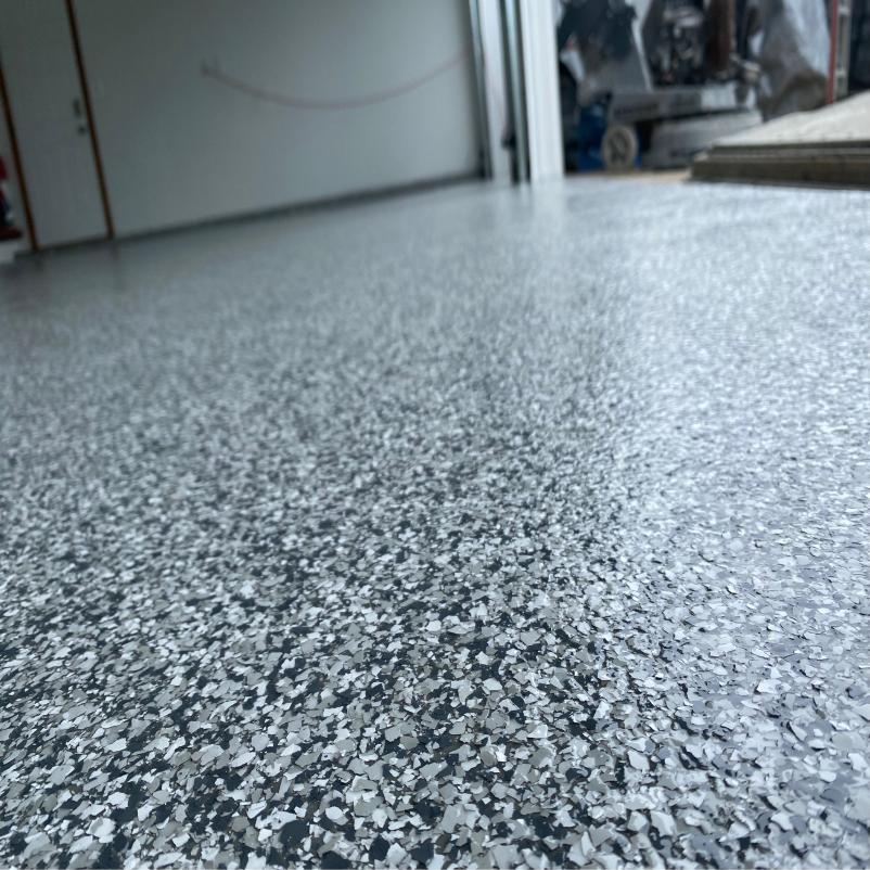 A view of a speckled, epoxy-coated floor surface in a garage area with an open door revealing construction equipment and materials.