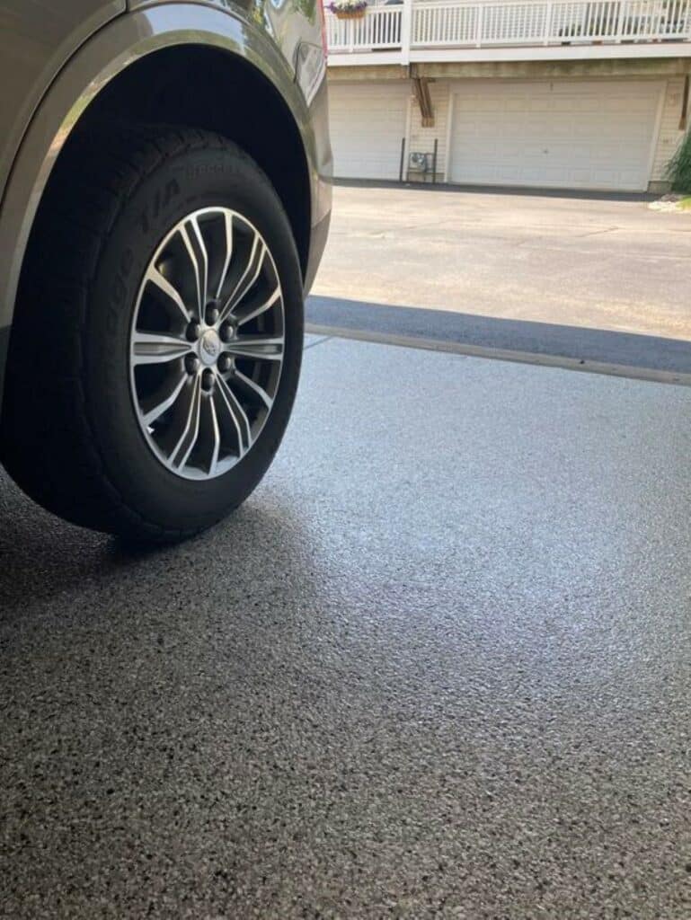 A vehicle’s tire is visible in a garage with a closed door. An adjacent building with a balcony and garages is in the background.