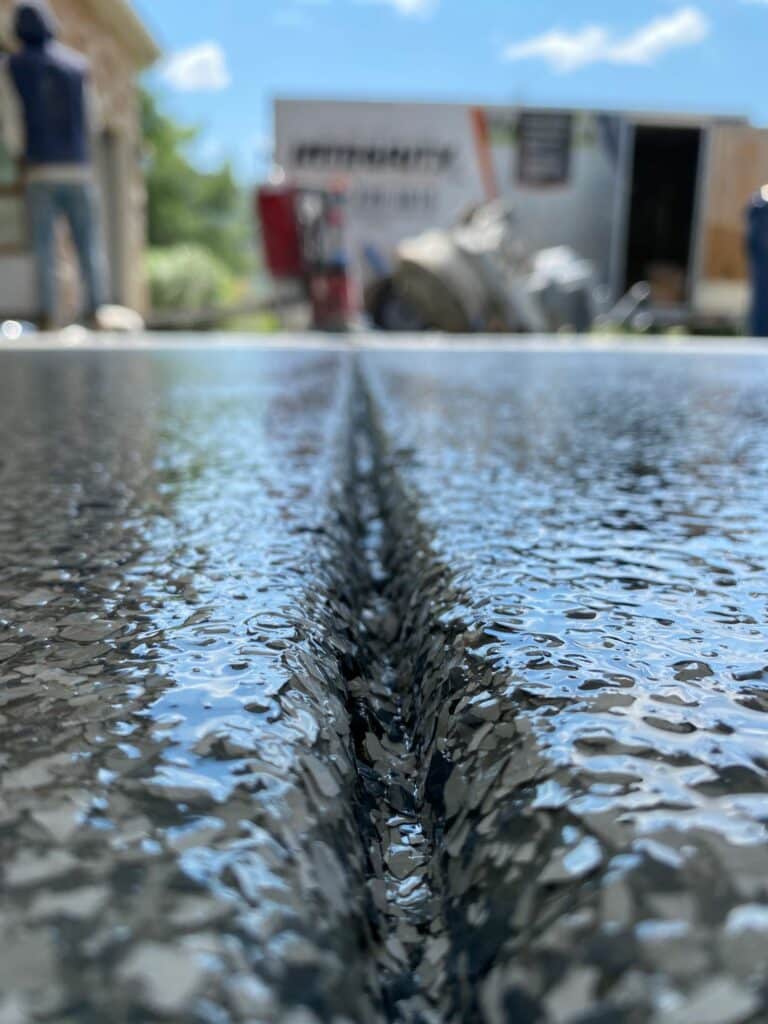 A close-up of wet pavement with construction activity in the background, featuring two persons and a partially visible trailer.