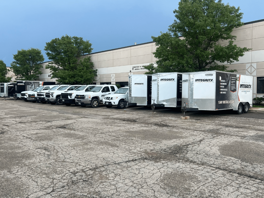 A parking lot with trucks, trailers, and a non-descript building in the background. Trees line the back, and cloudy sky overhead. No people visible.