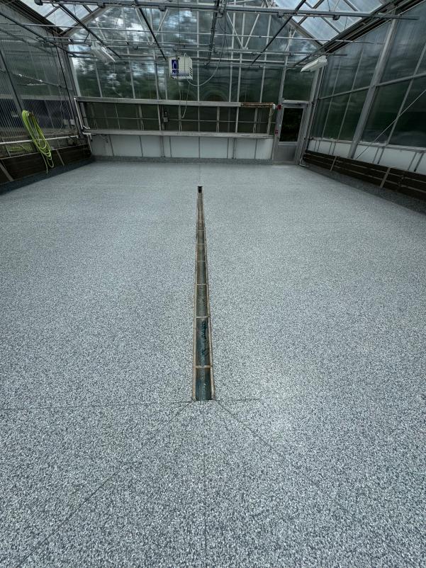 A greenhouse interior features a pebbled floor and HVAC system, with clear glass panels and metal framing, and a central drainage channel.