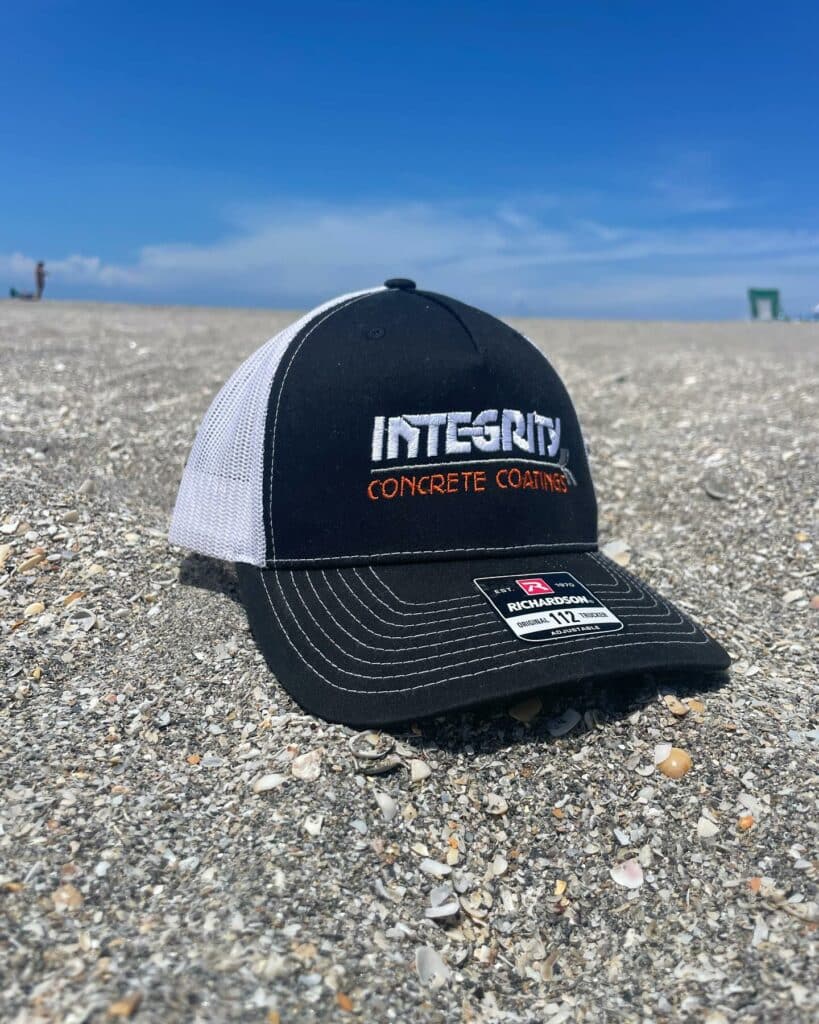 A cap with "Integrity Concrete Coatings" rests on a rocky beach with a clear blue sky; a person is visible in the distance.