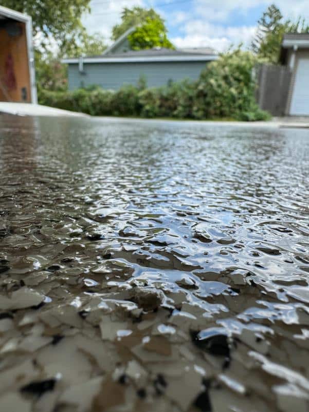 Close-up of a wet, textured surface with a green house, garage, and trees blurred in the background under a blue sky.