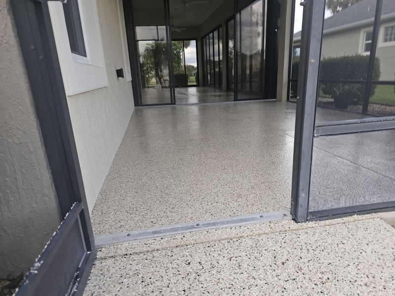 A screened-in porch with smooth, speckled flooring leads to the entrance of a house; greenery and lawn are visible outside.