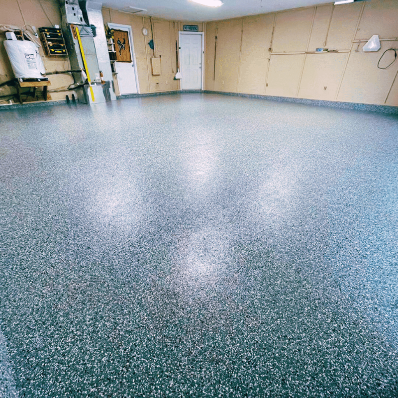 The image depicts an empty garage with a speckled epoxy floor, beige walls, a water heater, and tools mounted on the wall.