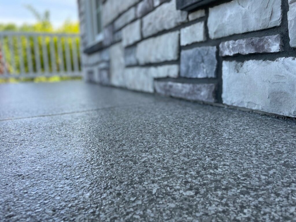 Close-up view of a textured concrete surface next to a stone wall, with a white railing and greenery in the blurred background.
