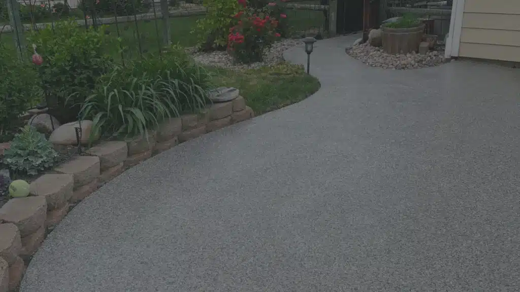 A landscaped backyard features a curved concrete pathway, vibrant garden beds with blooming flowers, decorative rocks, and small potted plants along the path's edge.