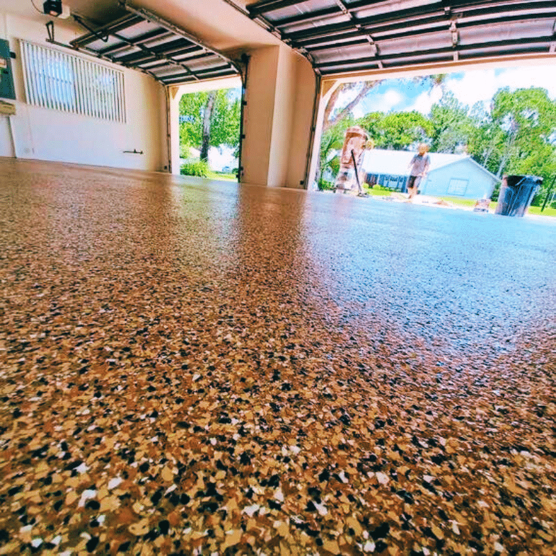A garage with a polished, speckled concrete floor. Two people work outside on a sunny day. Trees and a house in the background.