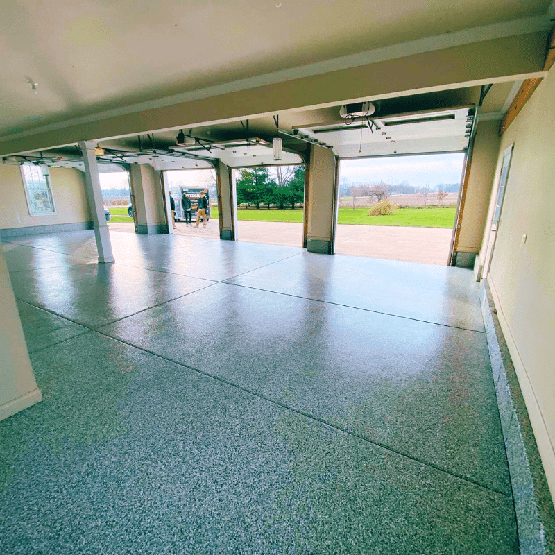 Spacious garage with shiny epoxy floor, open doors revealing a person outside. Green landscape and van parked on the pavement.