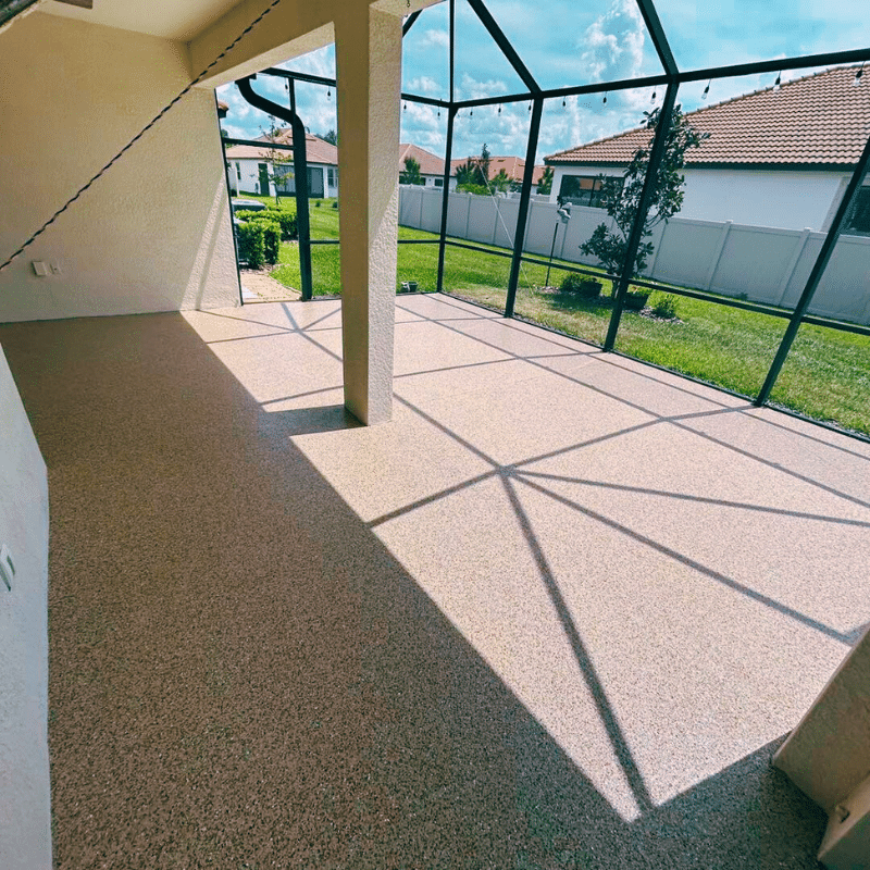 Enclosed patio with textured flooring and metal frame, overlooking grass yard and neighboring houses under a clear blue sky.
