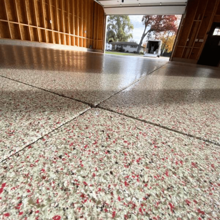 A garage with polished speckled flooring, unfinished wood walls, and an open door view of a driveway with trees and a residential house.
