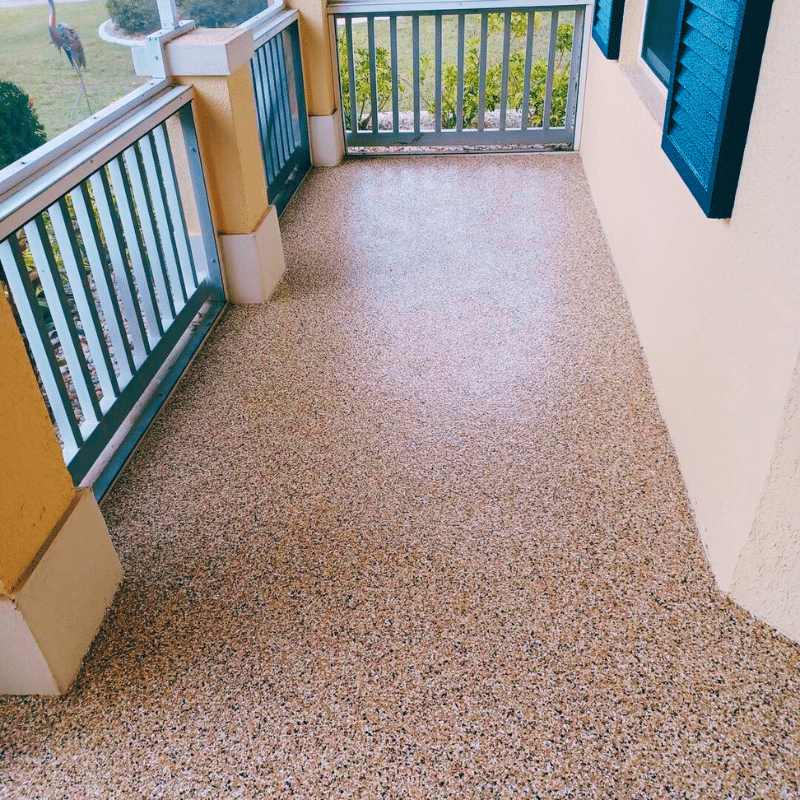 A clean, pebble-finished porch with blue shutters, enclosed by a wooden railing, overlooks a garden area with lush greenery on a bright day.