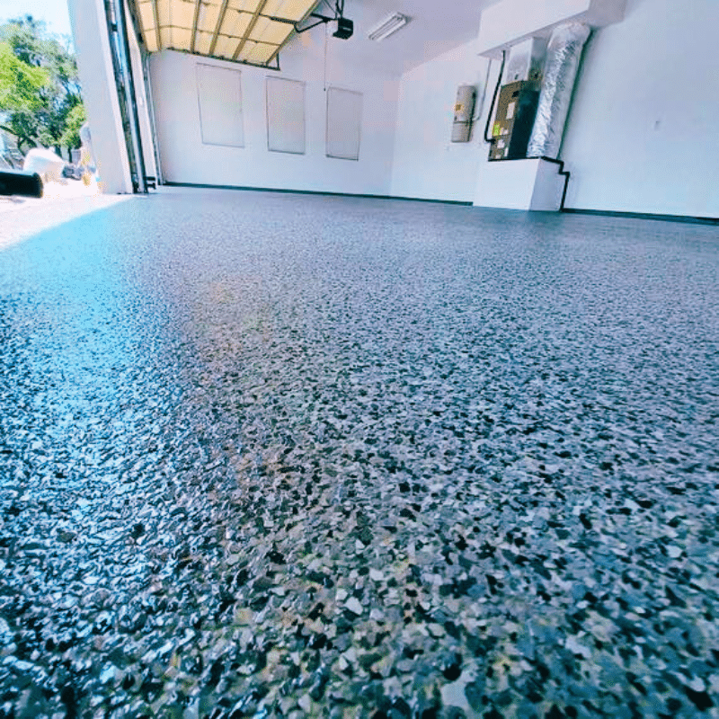 Smooth epoxy-coated garage floor, open door with trees visible outside. White walls and ceiling, featuring a utility area with electrical panels.