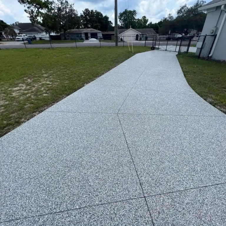A newly paved concrete path curves beside a grassy lawn, fenced yard, and residential houses in a suburban neighborhood.