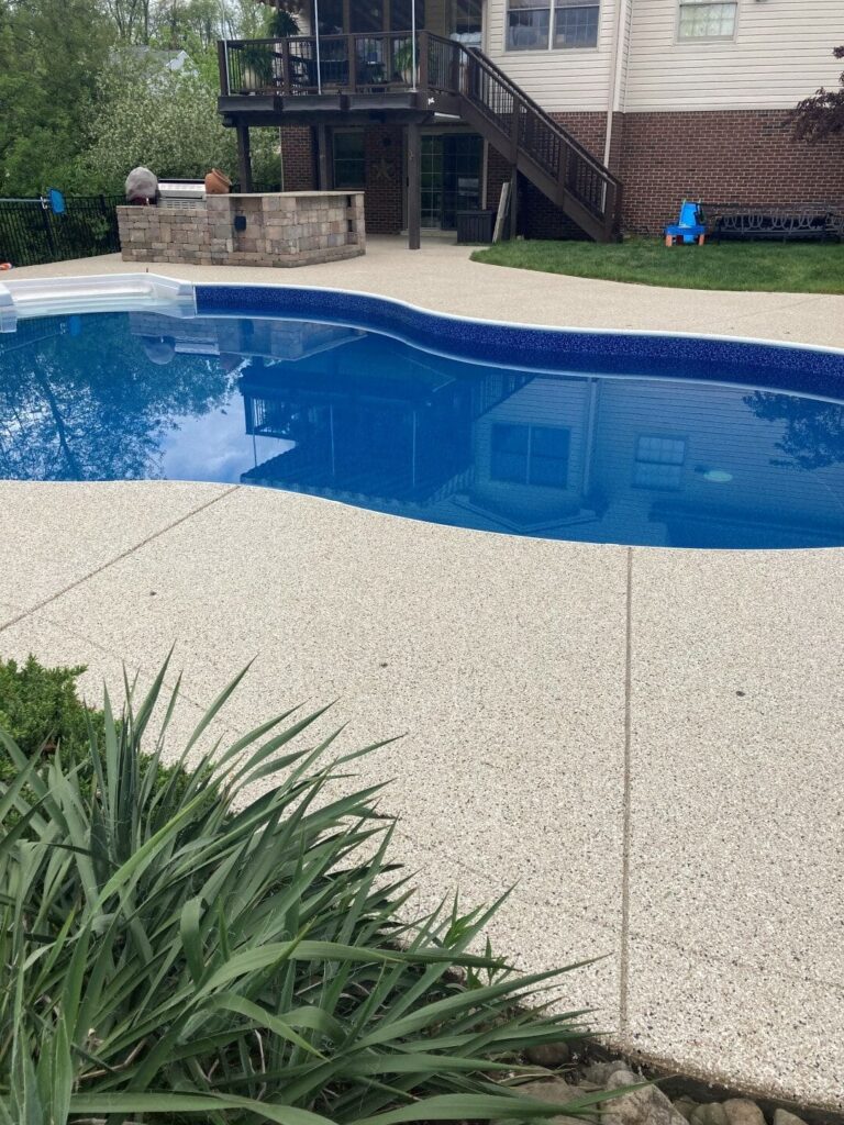 A backyard pool with clear blue water, surrounded by a concrete patio. Two people are near a brick outdoor structure, with grass nearby.