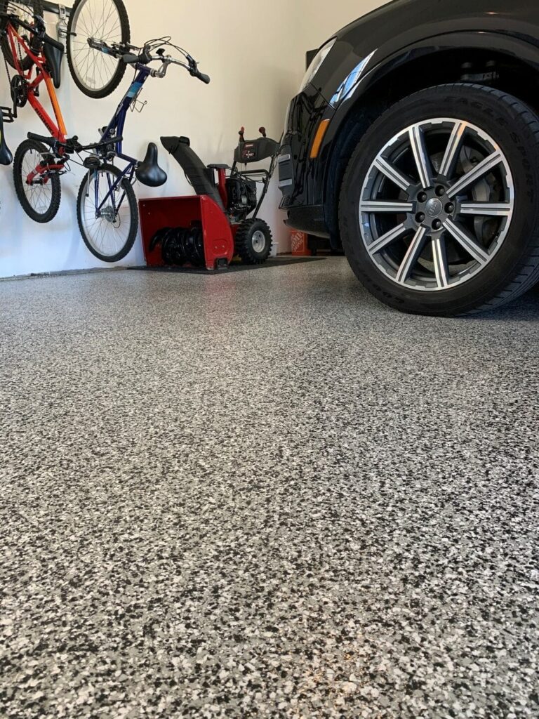 A garage with a speckled floor, bicycles, a car, and a snowblower. The space is clean and well-organized, with storage items.