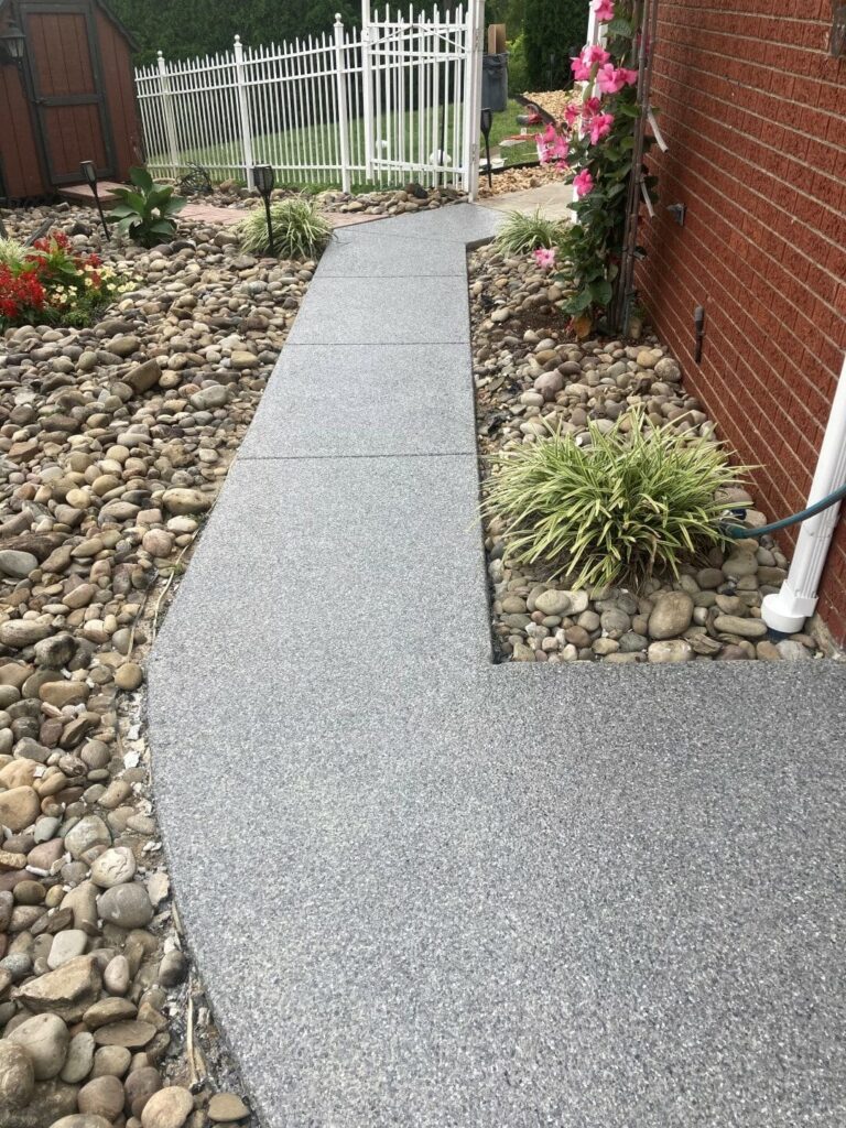 Modern stone pathway bordered by vibrant flowers and decorative rocks, leading to a white fence beside a red brick building.