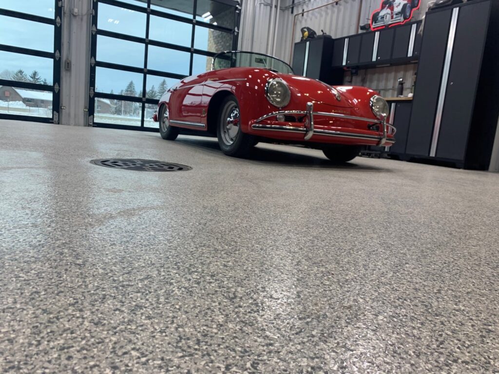A vintage red convertible is parked indoors on a polished floor. The garage has large windows and storage cabinets, showcasing a neat, organized space.