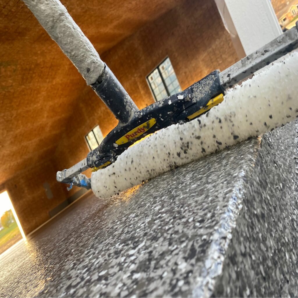 Close-up of a paint roller applying a speckled coating on a floor in a partially constructed room. Natural light filters through windows.